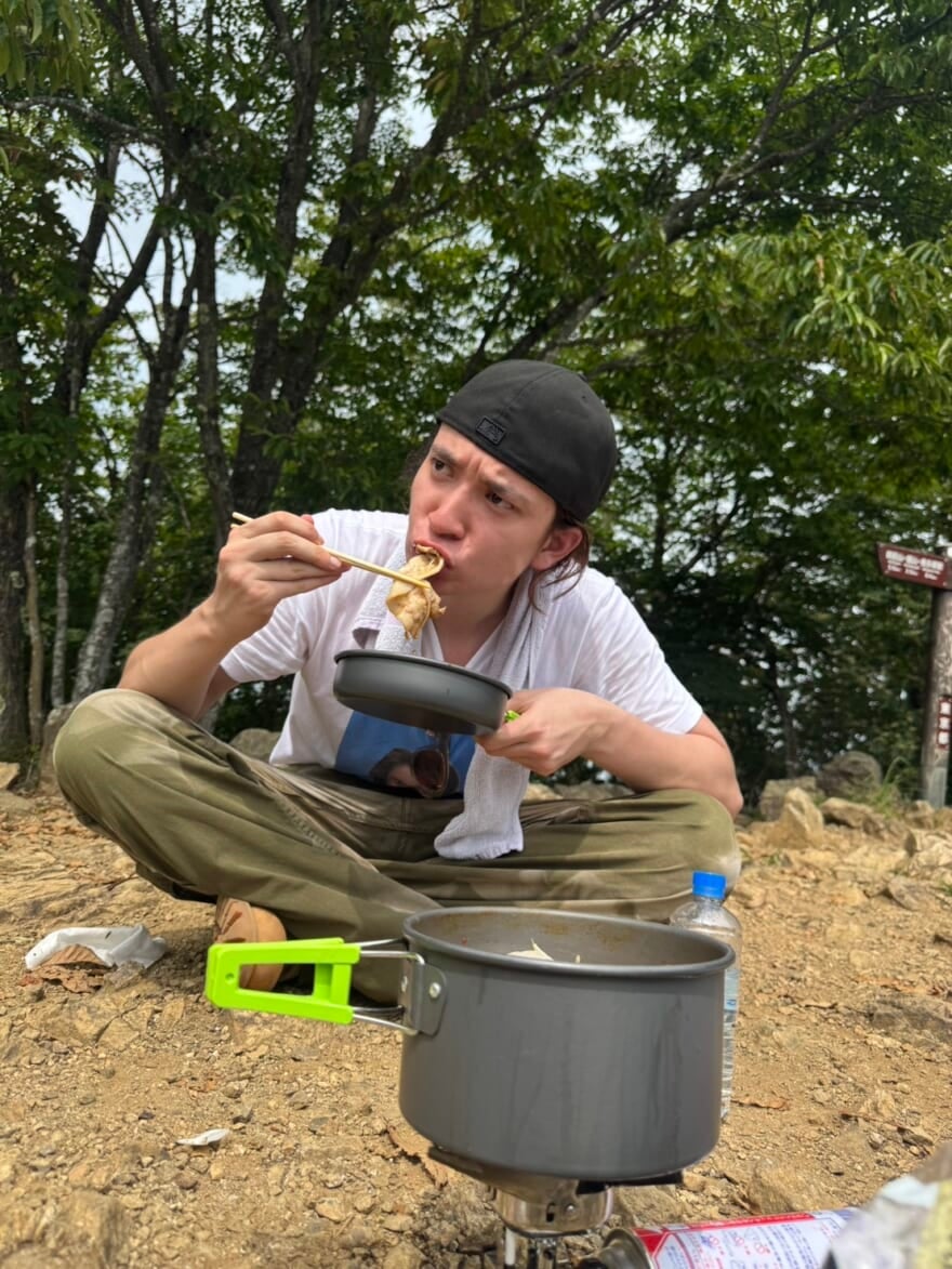 山頂で鍋を食べるメンズノンノモデル嵐翔真。自分の顔がプリントされたTシャツを着ている。