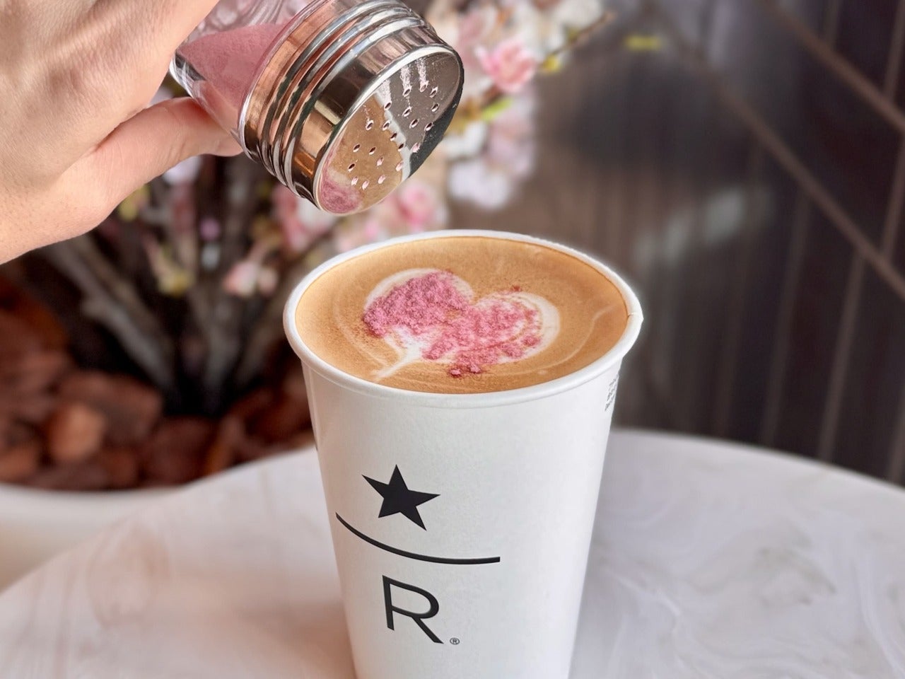 スタバ リザーブカフェ定番メニュー『クラシック ダブル トール ラテ』に、春限定コーヒーディスカバリーカウンター「サクラブルーミングパウダー」を振りかけている様子